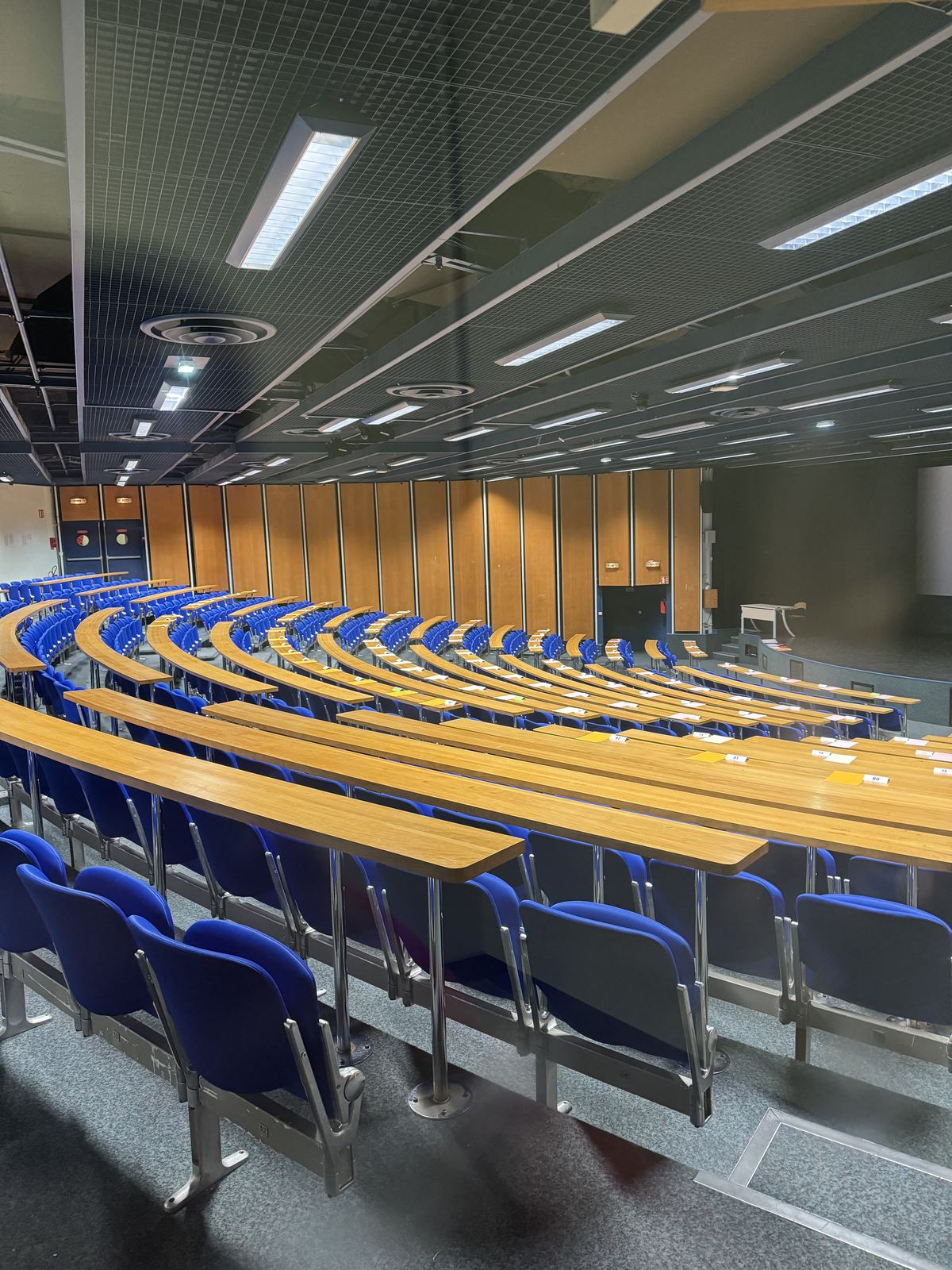 An empty tiered lecture hall with blue seats and long wooden desks