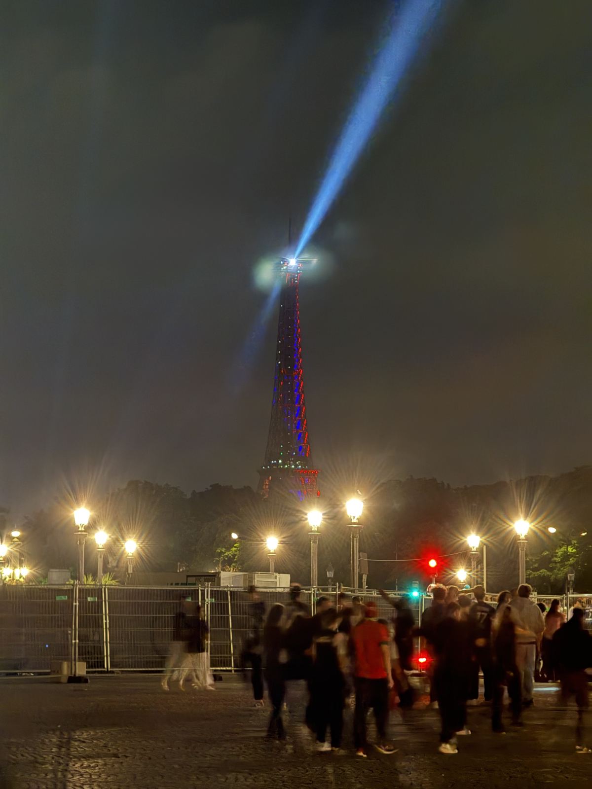 The Eiffel Tower lit at night, its beacon sweeping the sky above a crowded crossing